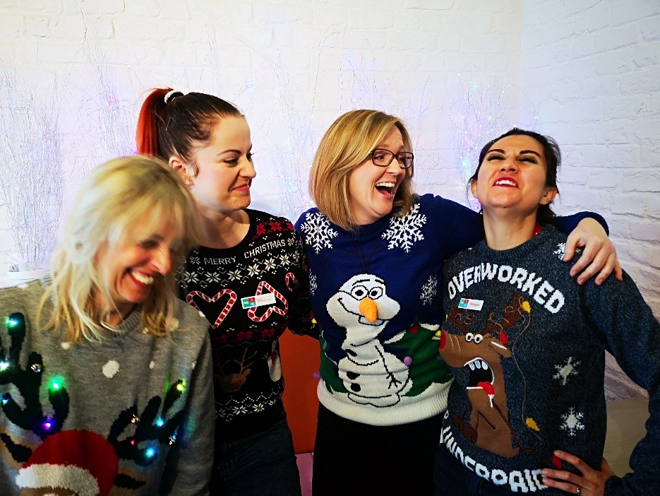 A group of four women smiling with their arms around each other. They're wearing Christmas jumpers.