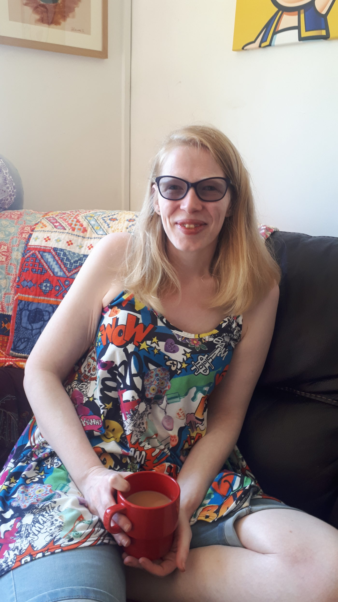 An image of Vicky. She is sat down wearing glasses and holding a cup of tea. She has long blonde hair and a floral vest top on.