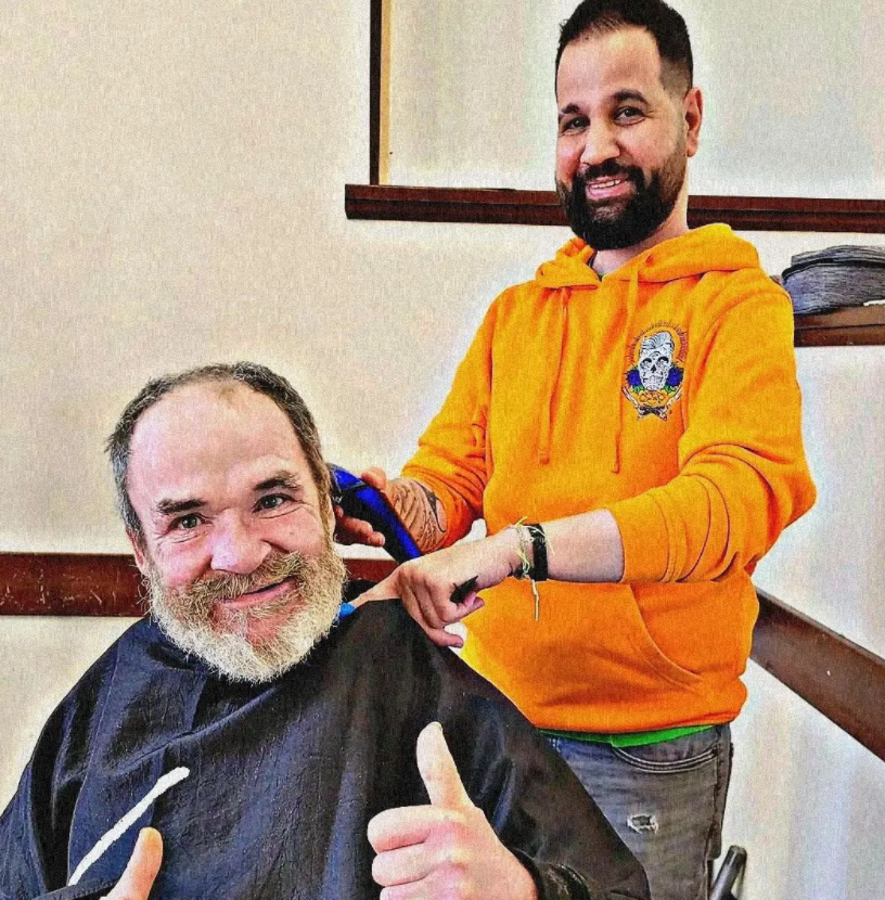 A barber is shaving a man's head. The barber is wearing an orange hoody and smiling, and his client is looking into the camera and smiling with his thumbs up. 
