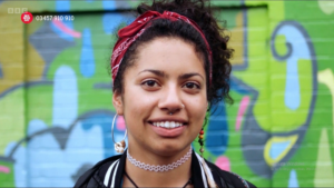 Close up of Dandi's face, the client from the video. She is wearing a red headscarf and smiling. There is a graffiti wall in the background. 